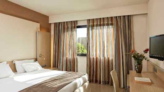 Ett hotellrum med en bäddad säng, skrivbord med stol, en TV och ett fönster med gardiner. (AI-genererad text)