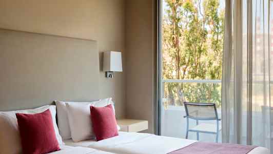 Hotellrum med två sängar, röda kuddar och utsikt mot en balkong med stolar och träd utanför. (AI-genererad text)