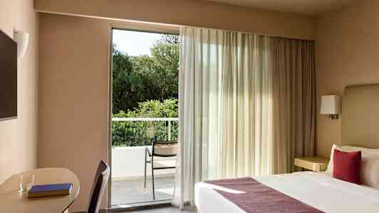 Ett hotellrum med en dubbelsäng och en arbetsyta bredvid. Rummet har ett stort fönster som leder ut till en balkong med en stol och utsikt över grönska. (AI-genererad text)