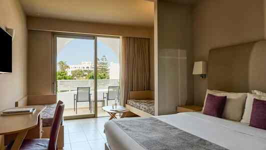 Ett modernt hotellrum med en dubbelsäng, skrivbord, sittmöbler och ett stort fönster som leder ut till en balkong med utsikt över en trädgård. (AI-genererad text)