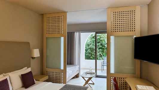 Hotellrum med dubbelsäng, sittmöbler och glasdörr som leder till en balkong med utsikt över grönska. (AI-genererad text)