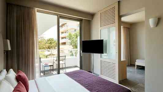 Ett hotellrum med en säng, en TV, en balkong med stolar och bord samt en utsikt över grönska och ett hus. (AI-genererad text)