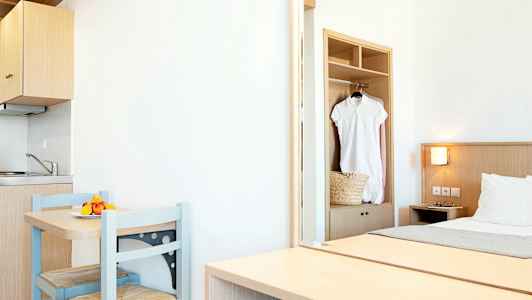 Ett ljust hotellrum med en liten köksdel, ett bord med två stolar och en säng. En vit klänning hänger i garderoben. (AI-genererad text)