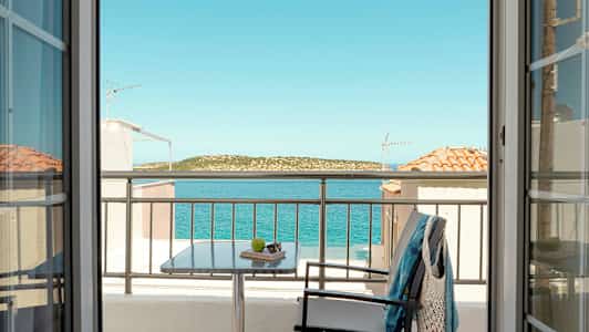 Utsikt från ett rum genom dörrar mot en balkong med bord, stol och klädesplagg, samt hav och landskap i bakgrunden. (AI-genererad text)
