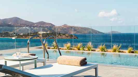 En utomhuspool med utsikt över havet och berg, omgiven av glasräcke och solstolar. (AI-genererad text)
