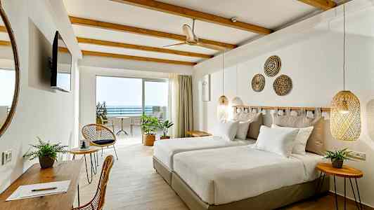 Ett hotellrum med två enkelsängar, väggdekorationer och möbler i ljusa färger. Utsikt mot en uteplats och havet. (AI-genererad text)