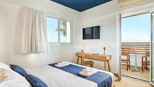 Hotellrum med säng klädd i blå och vit bäddning, skrivbord med stol, TV på väggen, och balkong med utsikt över havet. (AI-genererad text)