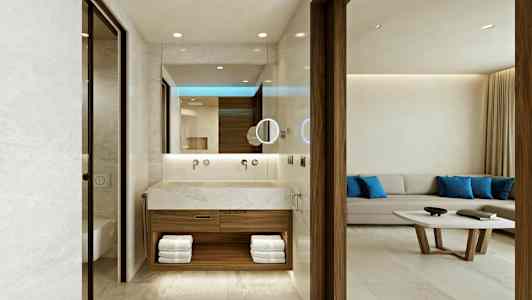 Ett hotellrum med ett öppet badrum som har en modern design med en handfat av sten och handdukar uppställda i hyllor. I bakgrunden finns en ljus soffgrupp med blå kuddar och ett soffbord. (AI-genererad text)