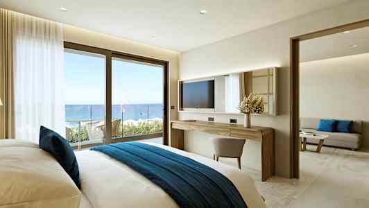 Ett hotellrum med en bekväm säng med blå sängöverkast, ett skrivbord med spegel, och en skjutdörr som leder till en balkong med havsutsikt. (AI-genererad text)