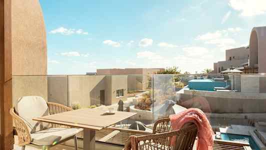 Vy från en terrass med glasräcke. Två korgstolar står vid ett litet bord. Utsikt över moderna byggnader, en pool och terrassområden under klarblå himmel. (AI-genererad text)
