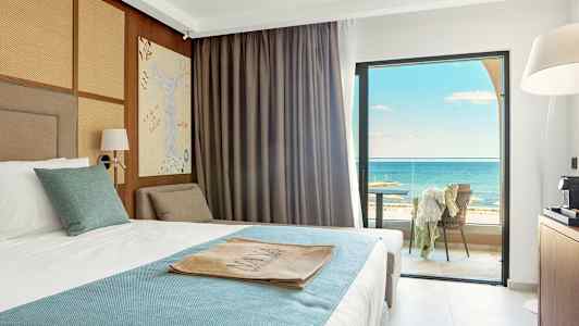 Hotellrum med en bäddad säng klädd i vit och blå sängklädsel. Rummet har stora fönster med utsikt över havet och en möblerad balkong. (AI-genererad text)