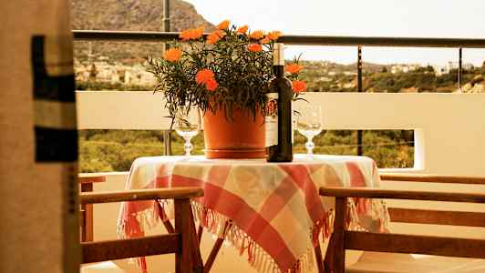 En terrass med ett bord klädd i en rutig duk, en krukväxt, ett vinflaska och två glas. Bakgrunden visar en naturskön utsikt. (AI-genererad text)