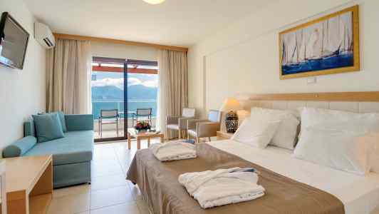 Ett hotellrum med en dubbelsäng, en soffa, ett bord med blommor och en utsikt mot havet genom glasdörrar. (AI-genererad text)