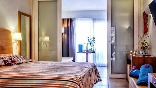 Ett hotellrum med en säng täckt av en randig överkast. I rummet finns även ett bord med dekorativa föremål och bakgrund med glasväggar. (AI-genererad text)