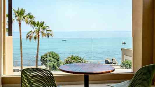 Utsikt från ett rum mot havet, med palmer och stolar på en terrass. (AI-genererad text)
