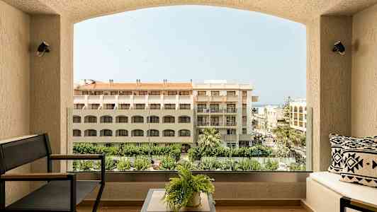 Utsikt från en balkong på ett hotell med soffmöbler, växter, samt byggnader och träd i bakgrunden. (AI-genererad text)