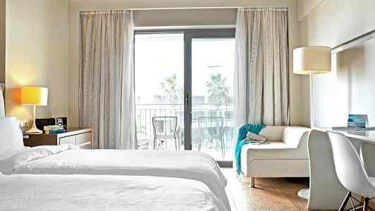 Ett hotellrum med vita sängkläder och en soffa framför en glasdörr till en balkong. (AI-genererad text)