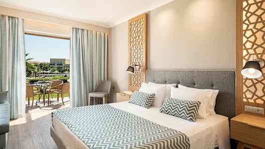 Ett hotellrum med en stor säng, dekorativa kuddar, och en öppning mot en balkong med utsikt över trädgård. (AI-genererad text)