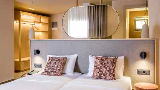 Ett hotellrum med två enkelsängar, dekorativa kuddar och en modern inredning. Bakom sängen finns en spegel och garderob i trä. (AI-genererad text)
