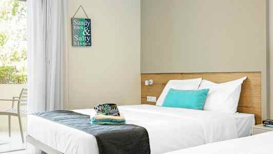 En hotellrum med två sängar och en vit sänggavel. På väggen hänger en tavla med texten "Sandy toes & Salty kisses." Ett öppet fönster ger utsikt mot grönska. (AI-genererad text)