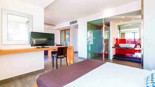 Ett modernt hotellrum med en säng i förgrunden, TV, skrivbord och en öppen badrumsdesign med en röd accentvägg. (AI-genererad text)