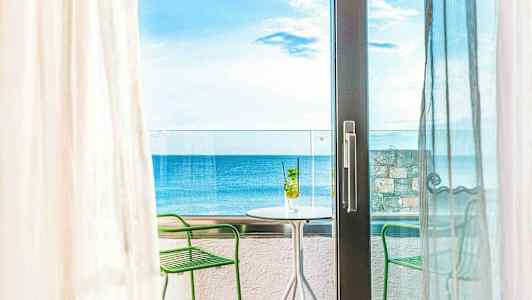 Utsikt från ett rum mot en balkong med bord och stolar, havet i bakgrunden. (AI-genererad text)