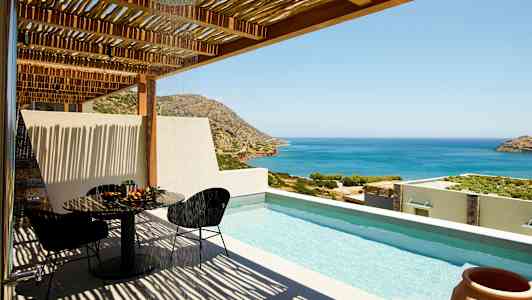 Utsikt över en terrass med pool, bord och stolar samt havet och bergen i bakgrunden. (AI-genererad text)