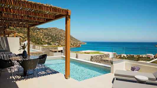 Bild av en terrass med en pool, solsängar och en matplats, med utsikt över havet och berg under en klar himmel. (AI-genererad text)