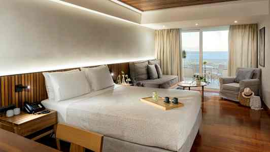 Ett hotellrum med en dubbelsäng, en sittgrupp med soffa och fåtölj, samt en balkong med utsikt över havet. (AI-genererad text)