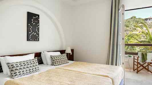 Rummet visar två enkelsängar med guldbeläckta täcken och en dekorativ kudde på varje säng. En väggprydnad hänger över huvudgaffeln, och ett fönster med utsikt över grönska och ett hus syns till höger. (AI-genererad text)