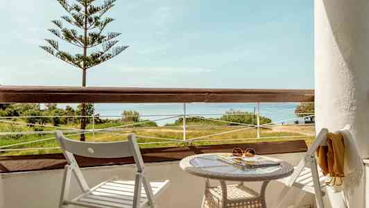 En balkong med utsikt över landskap och hav, med två vita stolar och ett bord. (AI-genererad text)