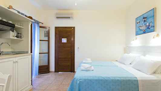 Ett hotellrum med två bäddar klädda i blå sängkläder, en träbeklädd dörr, ett mindre kök med diskho och en målning på väggen. (AI-genererad text)