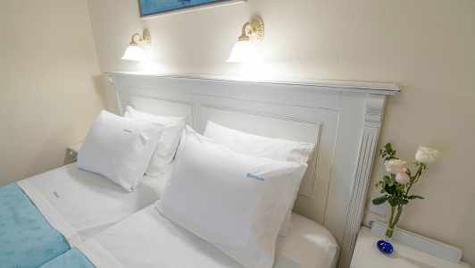 Ett uppbäddat dubbelsäng med vita lakan, en blå sängfilt, vita kuddar, en tavla ovanför och en liten vas med blommor på ett nattduksbord. (AI-genererad text)