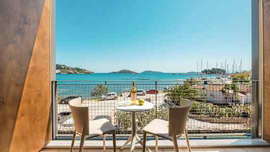 Utsikt från en balkong med två stolar och ett bord, mot havet och en småbåtshamn. (AI-genererad text)