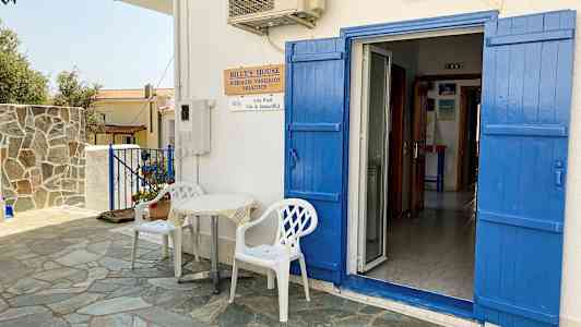 En vit byggnad med blå dörrar och fönsterluckor. Framför dörren står två vita plaststolar och ett litet bord. (AI-genererad text)