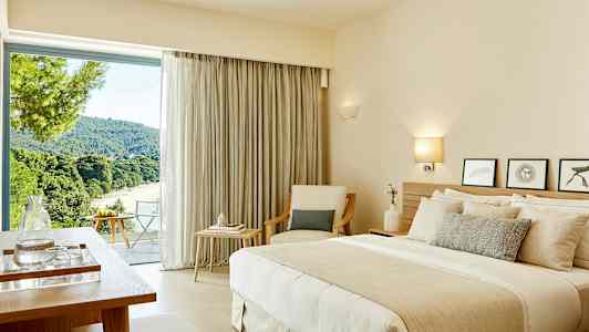 Ett hotellrum med en dubbelsäng, sittmöbler och ett stort fönster som erbjuder utsikt över en grönskande landskap. (AI-genererad text)