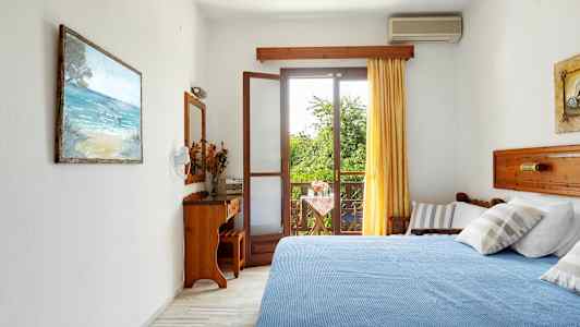 Ett hotellrum med en blå bädd, ett öppet fönster mot en balkong och gröna träd utanför. (AI-genererad text)