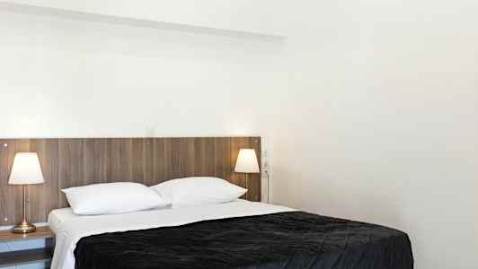 Ett sovrum med en dubbelsäng, vita sängkläder och en mörk överkast. Två sänglampor finns på sängbord bredvid sängen. (AI-genererad text)