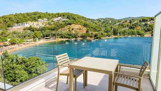 Utsikt från en terrass med stolar och bord mot en vik med klart blått vatten, omgiven av gröna kullar och byggnader. (AI-genererad text)