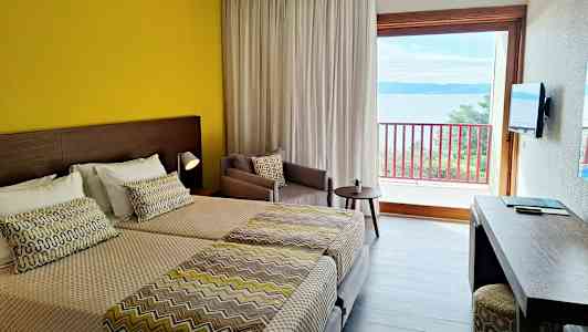 Ett hotellrum med två sängar, en gul vägg, ett skrivbord, en beige fåtölj och en balkong med utsikt över havet. (AI-genererad text)