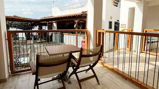 En uteplats med två trästolar och ett bord. Utsikt över en byggnad och ett tak. I bakgrunden syns en del av havet. (AI-genererad text)