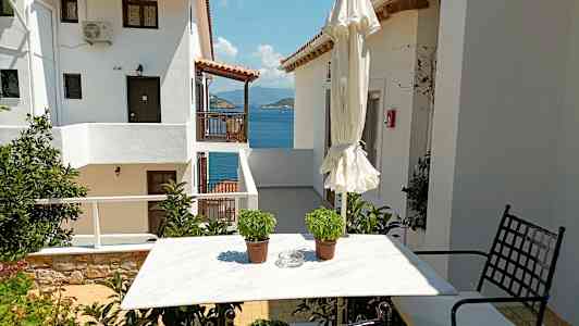 En vit husbyggnad med utsikt över havet, ett trädgårdsbord med två krukor och en hopfälld parasoll. (AI-genererad text)