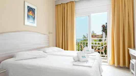 Ett ljust hotellrum med två enkelsängar, stora fönster med gulddraperier och en utgång till en balkong. (AI-genererad text)