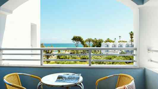 Utsikt från en balkong med havet i bakgrunden. En gul stol och ett bord finns på balkongen. (AI-genererad text)