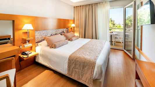 Ett hotellrum med dubbelsäng, sänglampor, ljusa träpaneler och en dörr som leder till en möblerad balkong. (AI-genererad text)