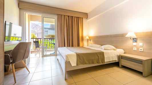 Ett hotellrum med dubbelsäng, skrivbord och stol. Balkongdörrar leder ut till en balkong med utsikt. (AI-genererad text)