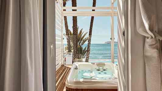 Utsikt från ett rum mot en terrass med bubbelpool, palmer och hav. (AI-genererad text)