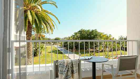 Utsikt från en balkong med ett bord, stolar och en handduk, mot en trädgård med palmer och gångväg. (AI-genererad text)
