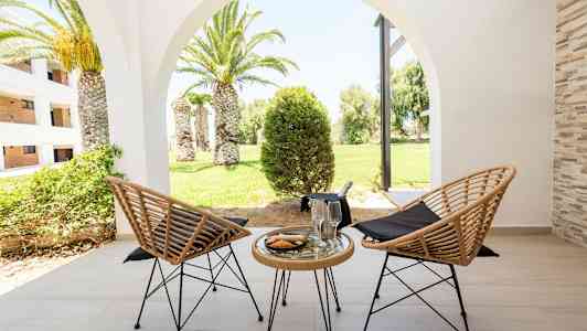 Två flätade stolar och ett bord med glas och en tallrik på en terrass, med utsikt över en grön trädgård med palmer. (AI-genererad text)