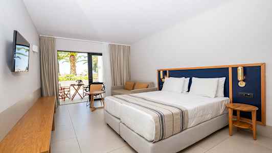 Hotelrum med en stor säng, sittgrupp och utsikt mot en terrass och trädgård. (AI-genererad text)